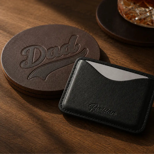A black leather card holder wallet with an embossed 'Parisian' logo, shown next to a dark brown circular coaster engraved with 'Dad' on a wooden table.