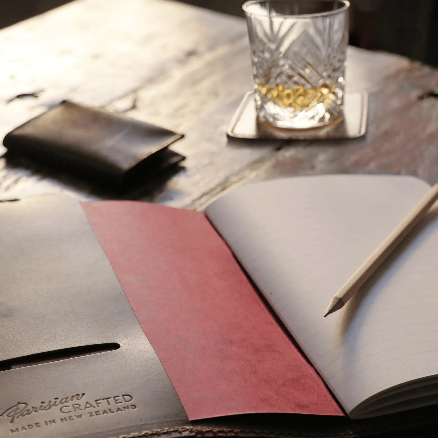 Open Parisian Crafted brown leather notebook on a rustic wooden table, paired with a handcrafted leather wallet, crystal glass of whiskey, and a pencil—capturing timeless elegance and refined Parisian craftsmanship.