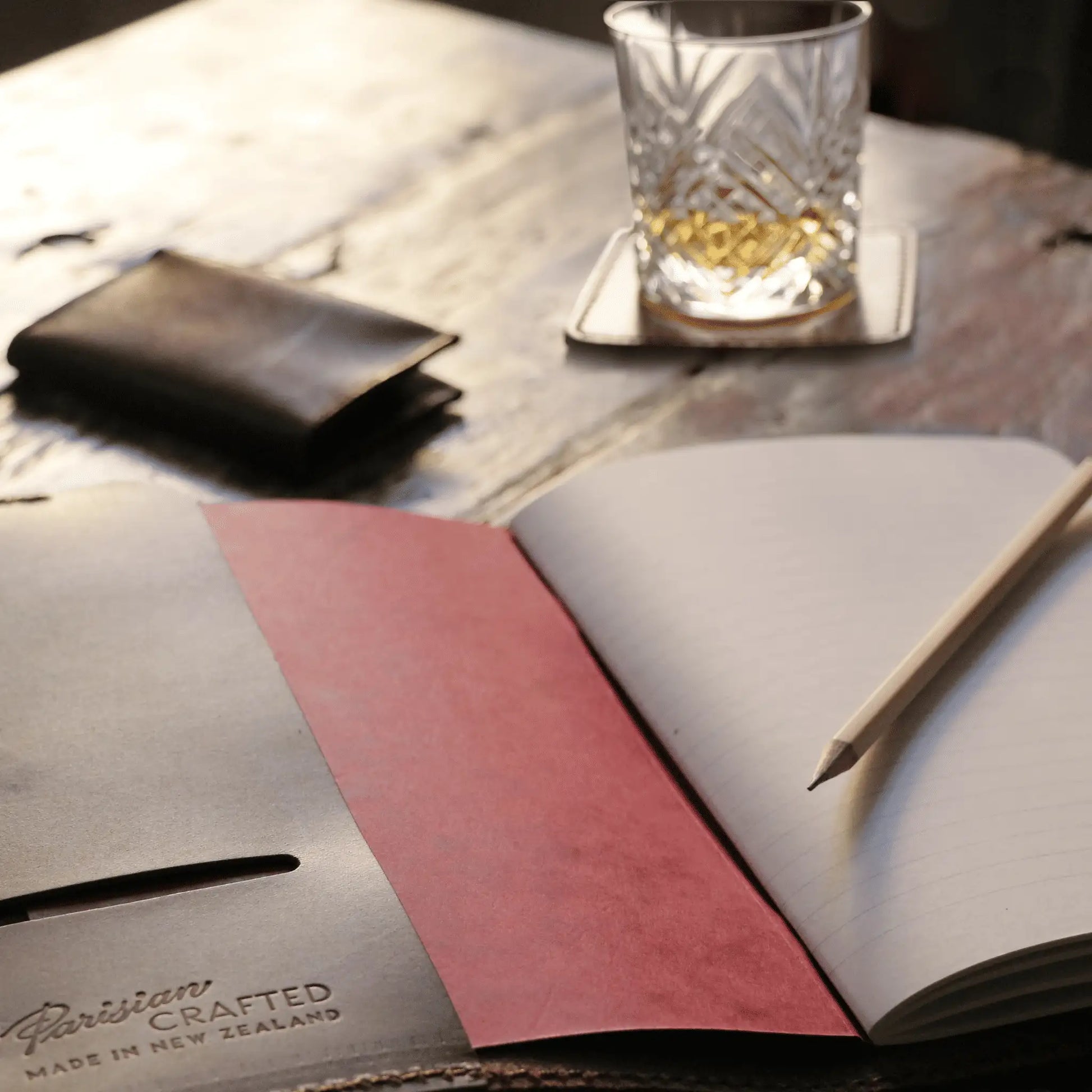 Open Parisian Crafted brown leather notebook on a rustic wooden table, paired with a handcrafted leather wallet, crystal glass of whiskey, and a pencil—capturing timeless elegance and refined Parisian craftsmanship.