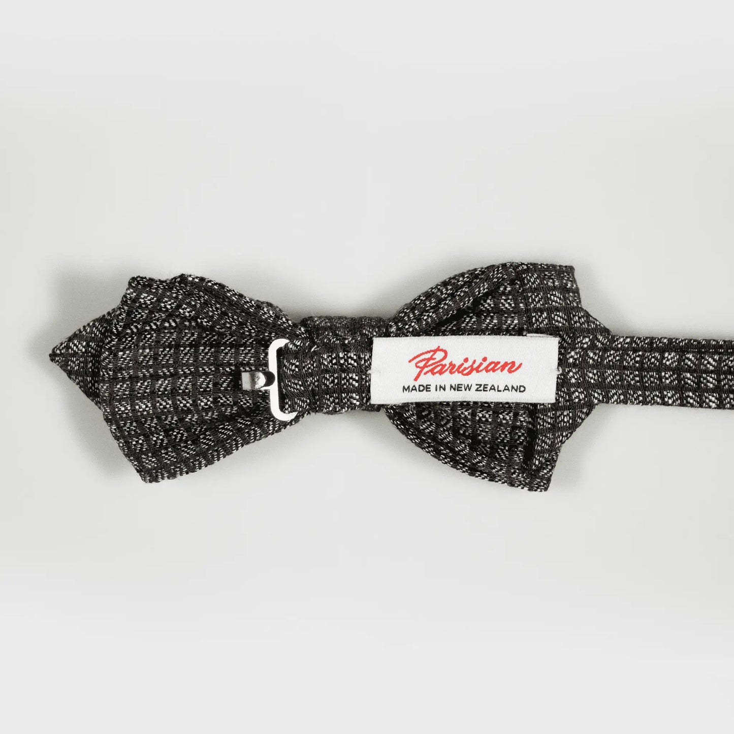 The back of a chocolate and grey micro-square patterned bow tie, displaying woven "Parisian Made in New Zealand" label.