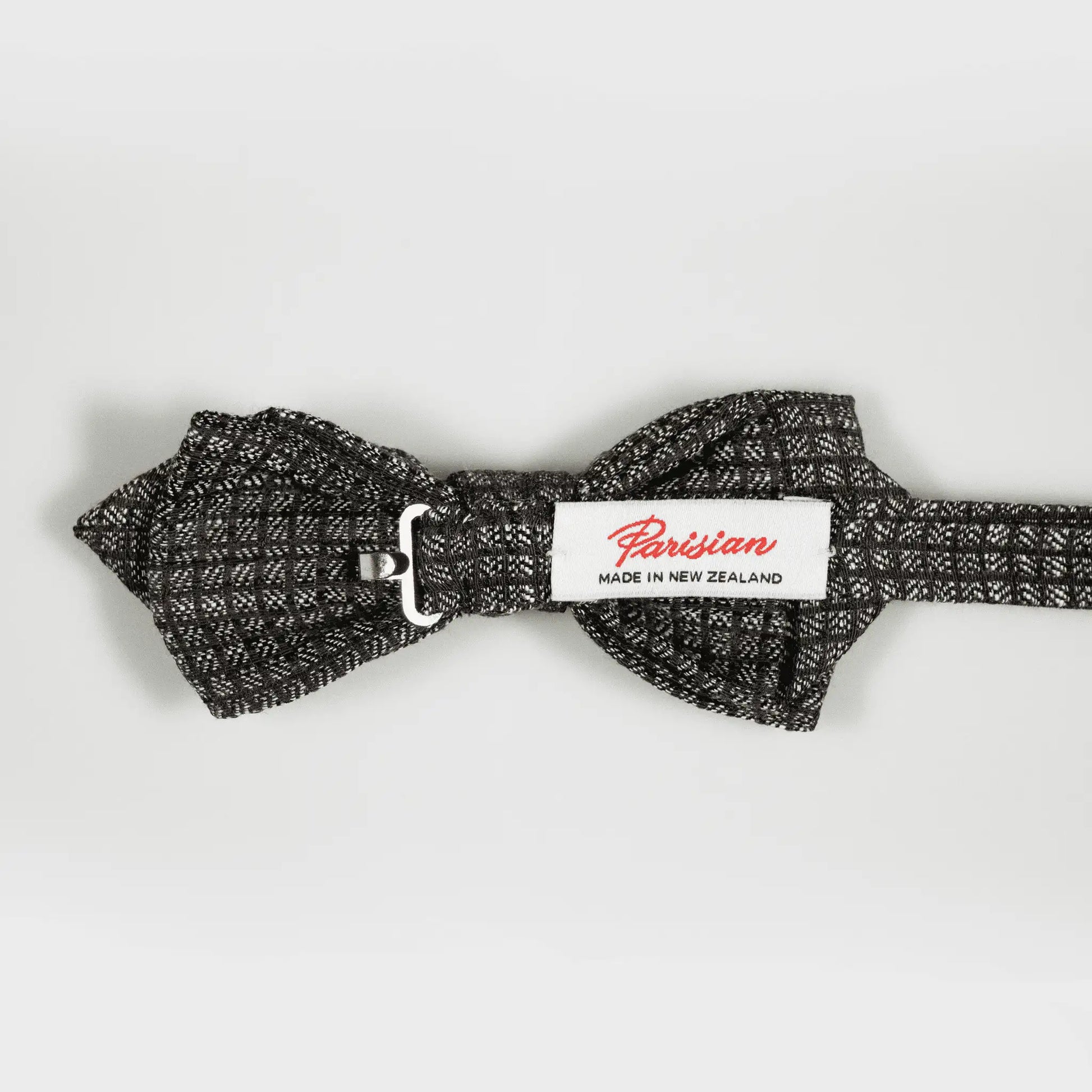The back of a chocolate and grey micro-square patterned bow tie, displaying woven "Parisian Made in New Zealand" label.