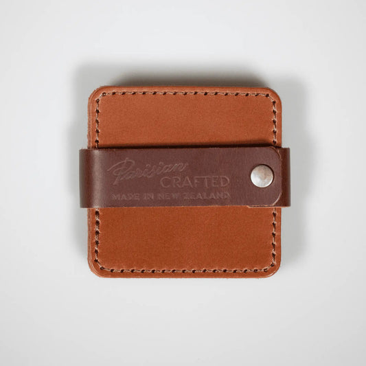 Set of four leather coasters in tan with leather strap and brass hardware. 'Parisian Crafted Made in New Zealand' embossed, showcasing premium craftsmanship.