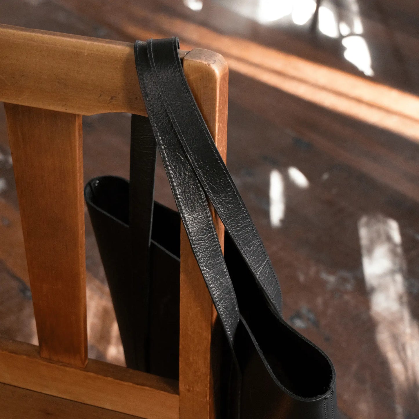 Black Leather Everyday Tote hanging on wooden chair, close-up showcasing quality leather and refined stitching—small batch, made to last, for men and women.