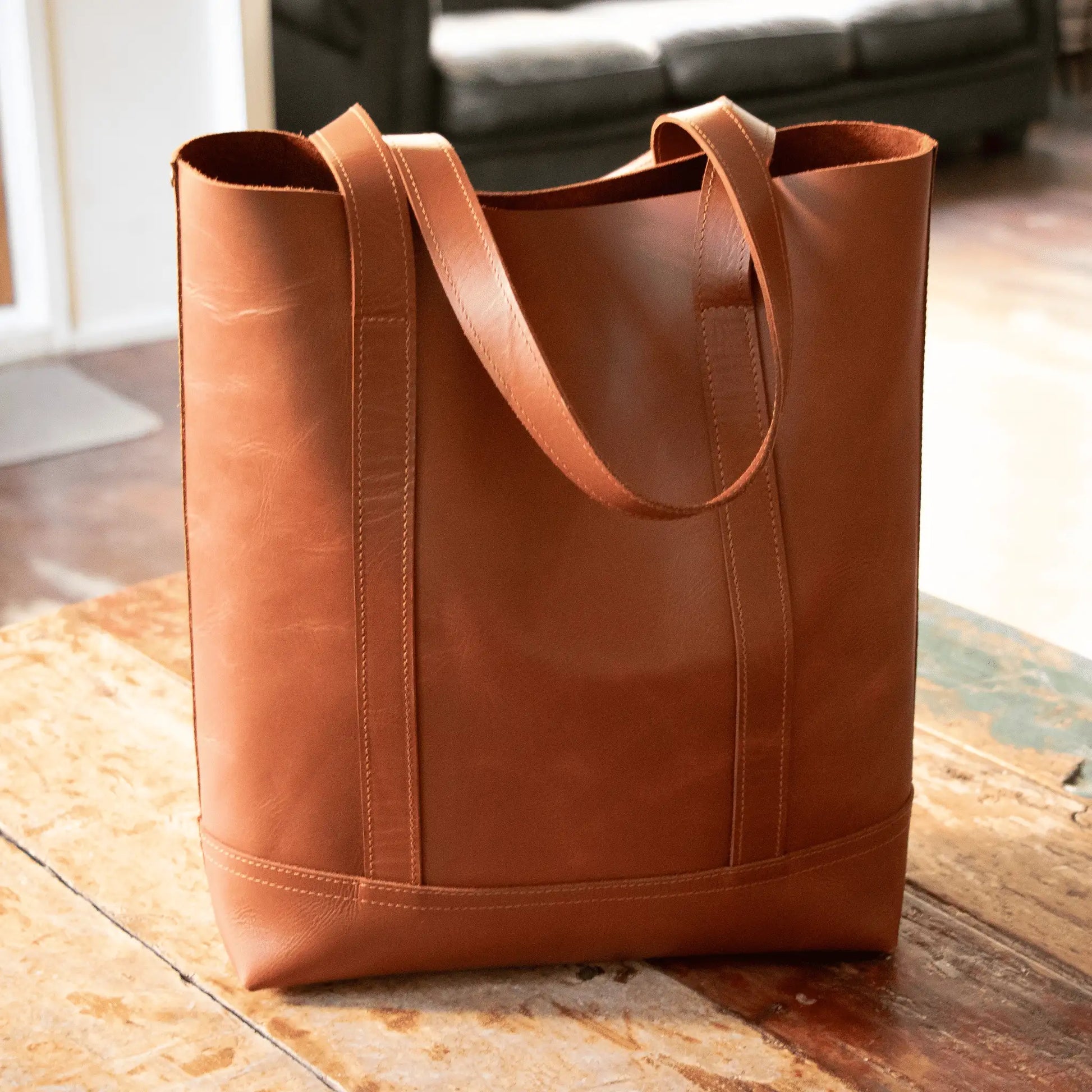 NZ-made tan leather tote on rustic wooden table in sunlight, showcasing expert craftsmanship and timeless, simple design.