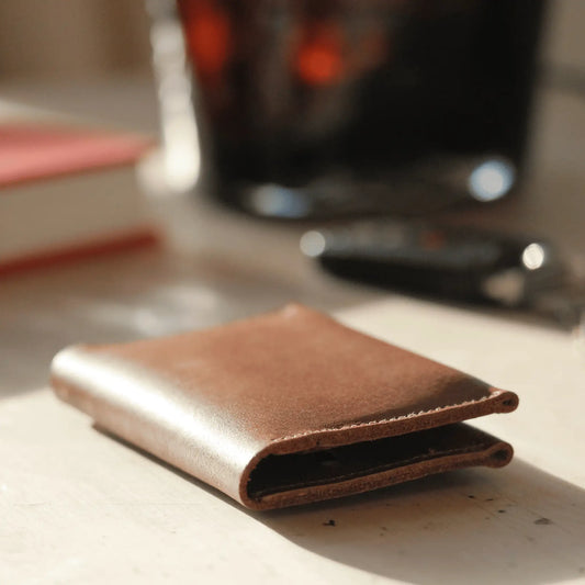 Close-up of tan leather folded card wallet sitting in the sun on a wooden table, showcasing its rich texture and quality craftsmanship.