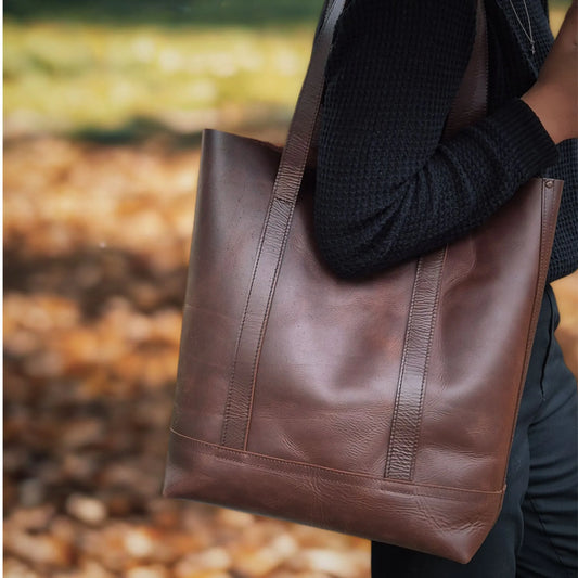 Person holding a chestnut brown leather tote bag outdoors with blurred background