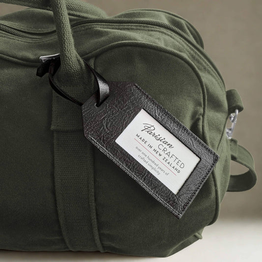 Green duffel bag with a 'Parisian Crafted' black leather luggage tag on a neutral background