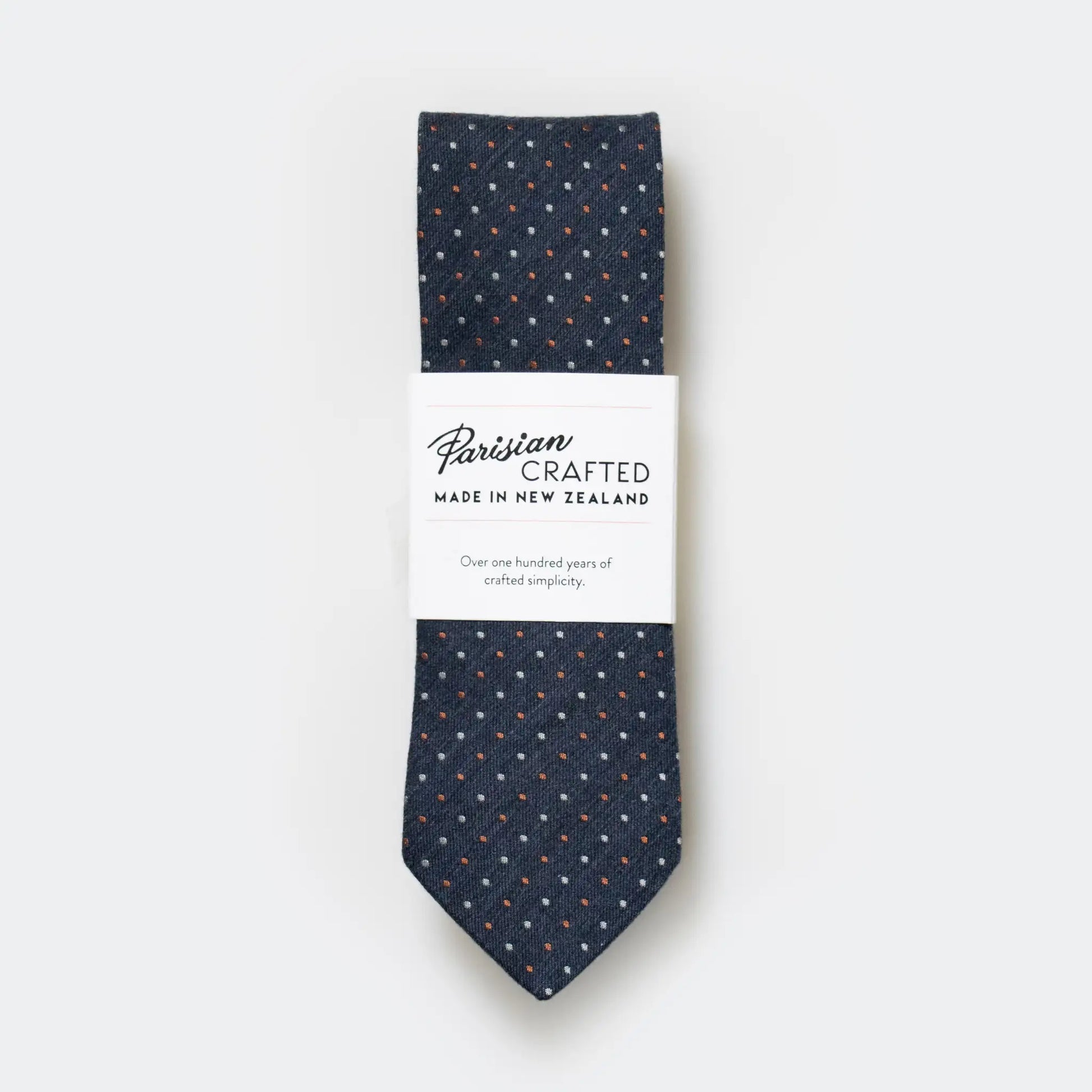Denim blue, white and rust pin-dot design necktie folded into Parisian Crafted's sleek packaging sleeve, on a white background.