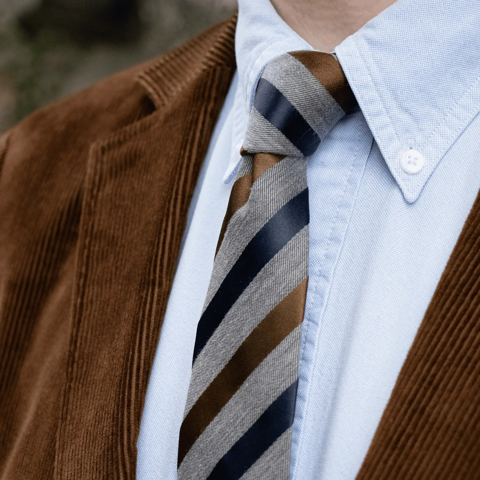 Close-up of the grey tie with navy and tobacco satin stripes, worn with a soft blue button-down shirt and brown corduroy blazer.