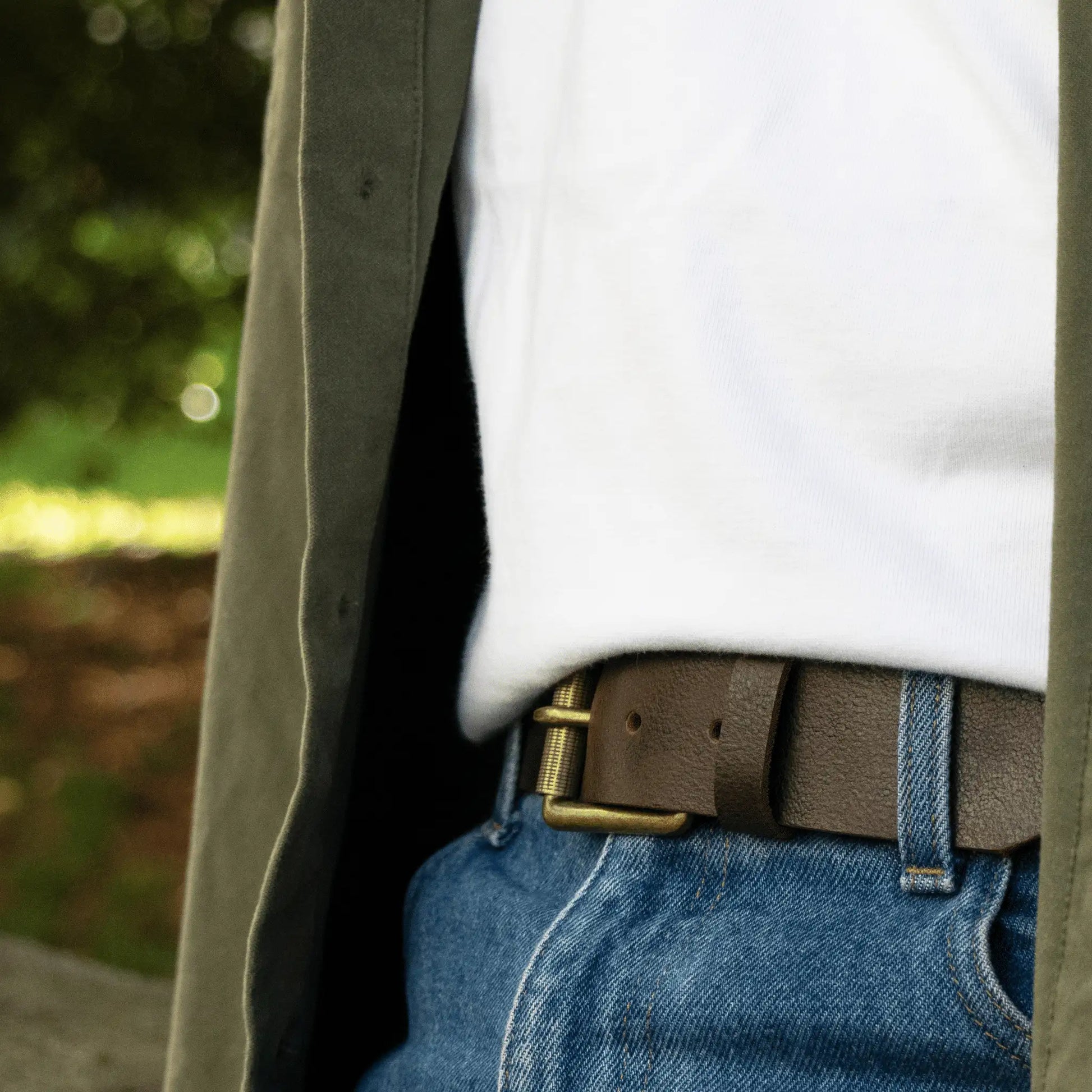 Rustic brown leather work belt worn with jeans, t-shirt and green shirt. Close-up to show brass buckle detailing.