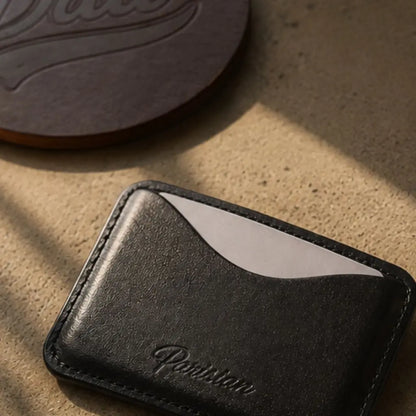 A close-up of a black leather card holder wallet with the brand name 'Parisian' embossed on the front, resting on a textured beige surface with warm sunlight and shadows.
