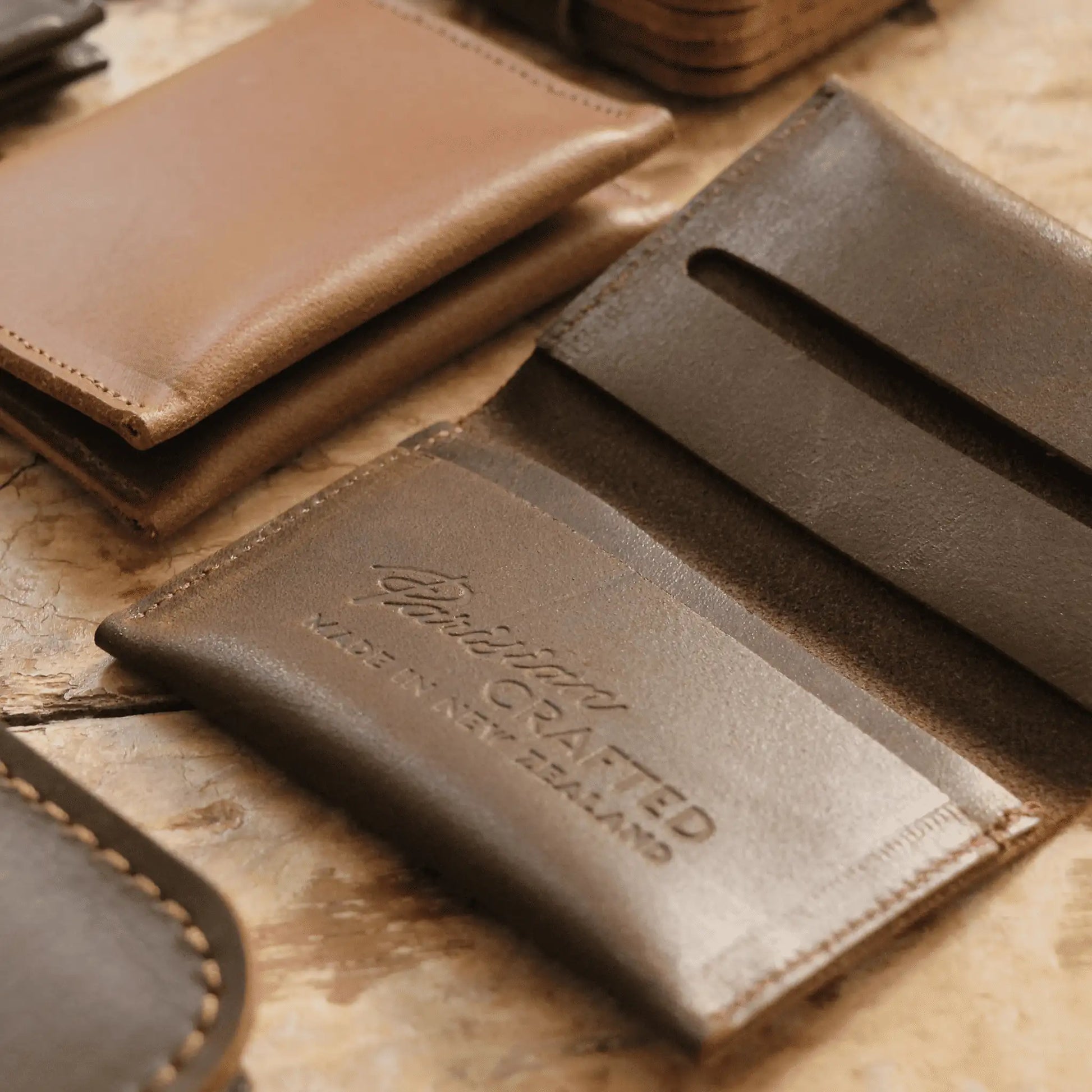 Close-up of an open brown leather folded card wallet showing the embossed "Parisian Crafted Made in New Zealand" text on a wooden table, surrounded by other leather wallets and goods.
