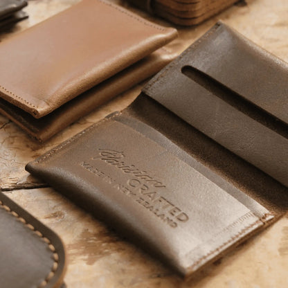 Close-up of an open brown leather folded card wallet showing the embossed "Parisian Crafted Made in New Zealand" text on a wooden table, surrounded by other leather wallets and goods.