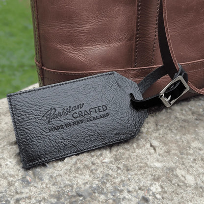 Black leather luggage tag with 'Parisian Crafted' branding on a brown leather bag.