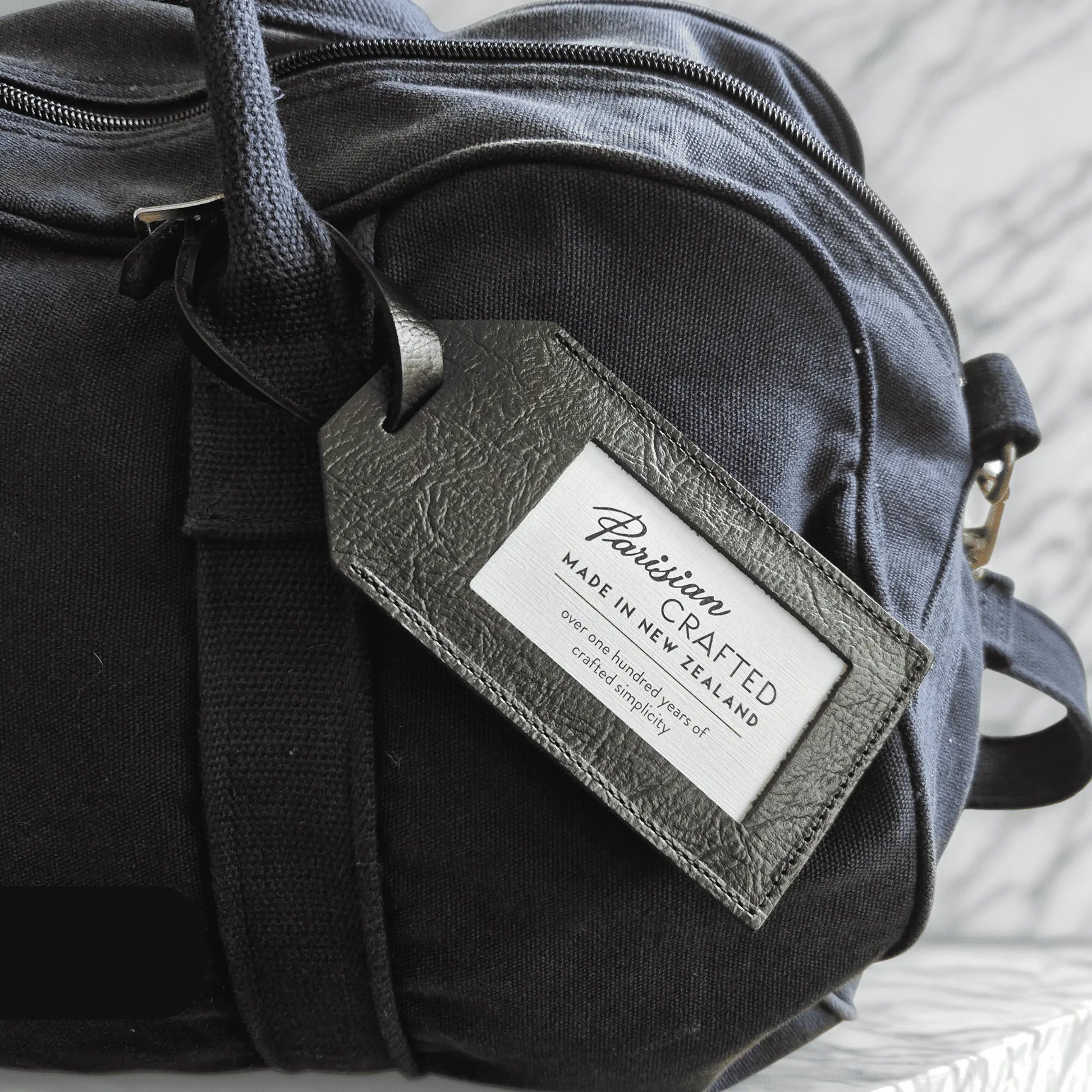 Navy duffel bag with a black leather tag displaying 'Parisian Crafted' on a light background