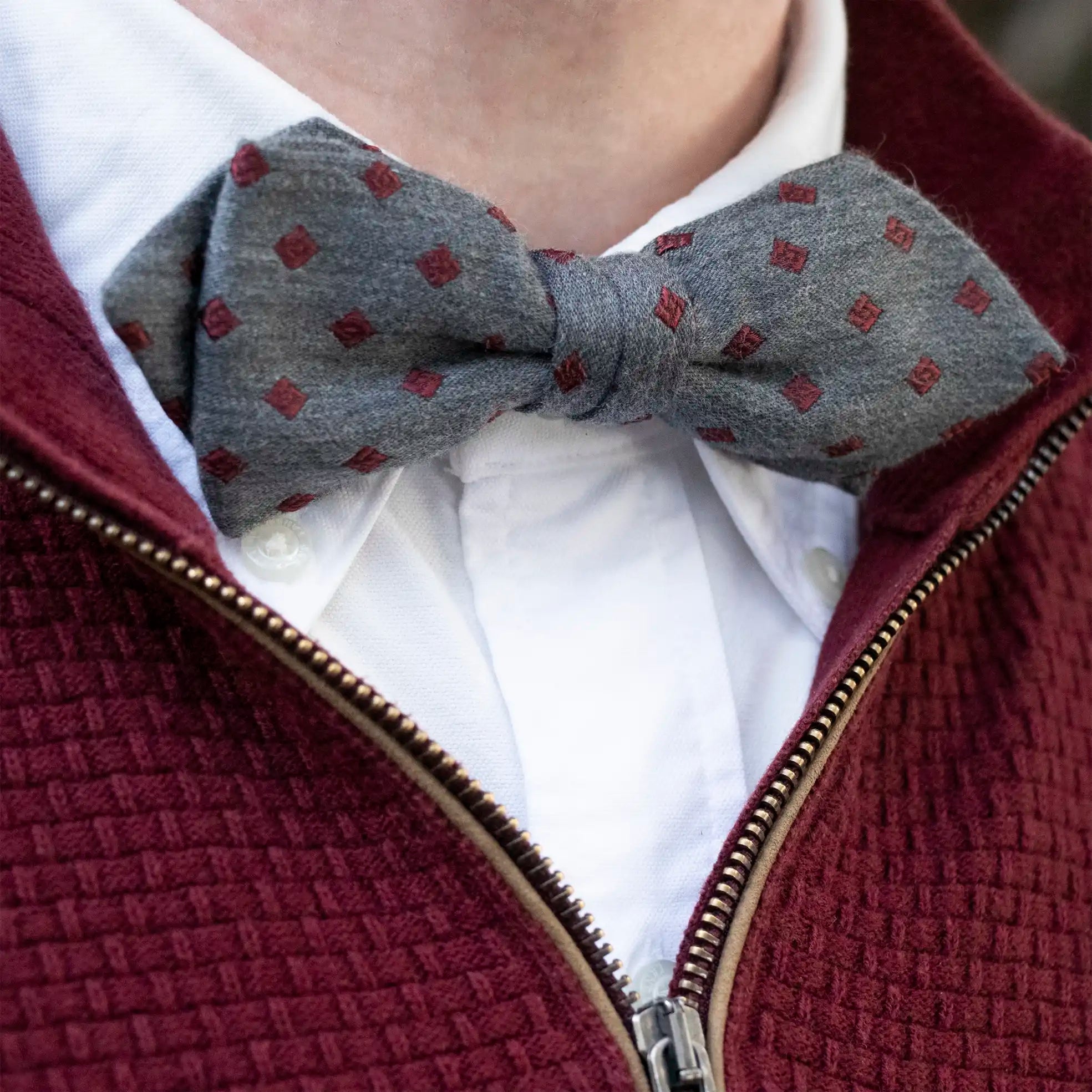 A close up of the  Grey Marle with Burgundy Diamond Bow Tie worn with a White Button Down Shirt and a Woven Burgundy Zip Up Jumper.