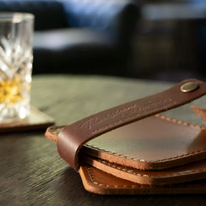 A close up of the Stitched Leather Coaster Set sitting on a wooden table, the leather strap is undone and there is a Crystal Glass of Whiskey in the background sitting on a Leather Coaster.