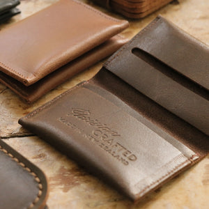 A close up of an open Brown Leather Folded Card Wallet showing the embossed text 'Parisian Crafted' on a wooden able surrounded by other leather wallets and goods