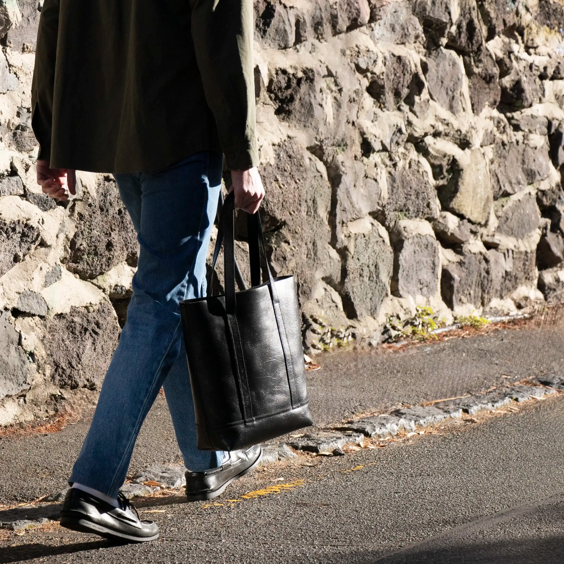 A Man carrying the Black Everyday Leather Tote walking in the sun up a street.